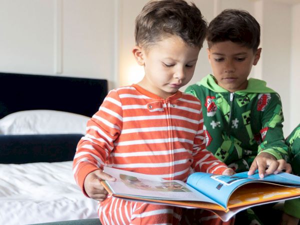 Two young boys in pajamas sit on a bed reading an illustrated book together in a bright room.