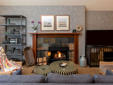 A cozy living room with a glowing fireplace, wooden mantel, flower vase, bookshelves, and pillows on patterned rugs.