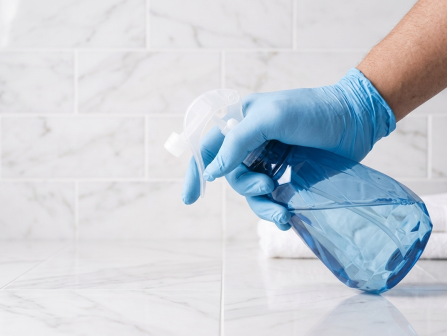A gloved hand sprays a blue liquid from a clear cleaning spray bottle on a white tiled floor.
