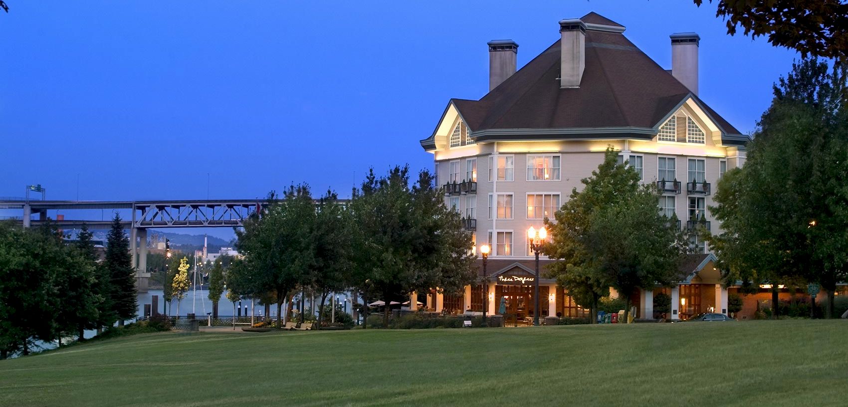 A riverside hotel building at dusk with a grassy lawn, trees, and a bridge in the background.