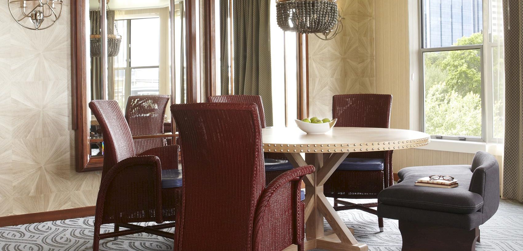 A bright dining room with a round table, cushioned chairs, beaded chandelier, patterned curtains, large windows, and a small bowl of limes.