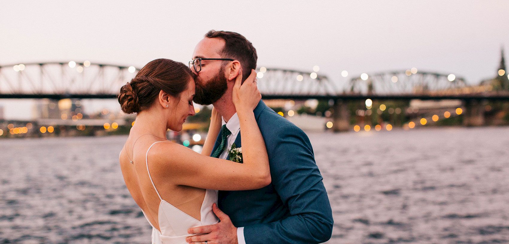 A bride and groom share a kiss on a beach at sunset with city lights glowing in the background.