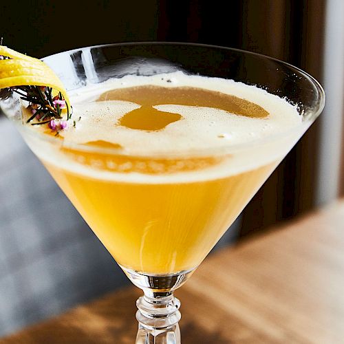 A bright yellow cocktail in a martini glass, topped with a banana slice and edible flowers, with frothy foam swirling on top.