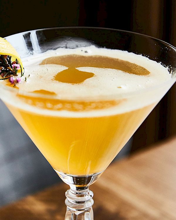 A bright yellow cocktail in a martini glass, topped with a banana slice and edible flowers, with frothy foam swirling on top.