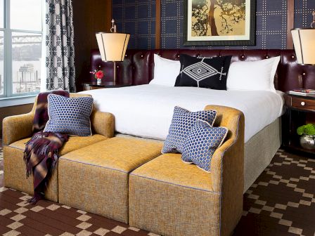 A chic hotel suite: king bed with patterned pillows, two warm lamps, and a yellow couch by window and curtains.
