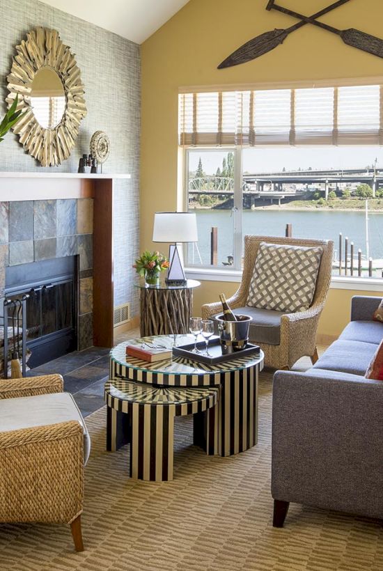 A cozy living room with a fireplace, wicker chairs, a striped coffee table, and a large window overlooking a river.