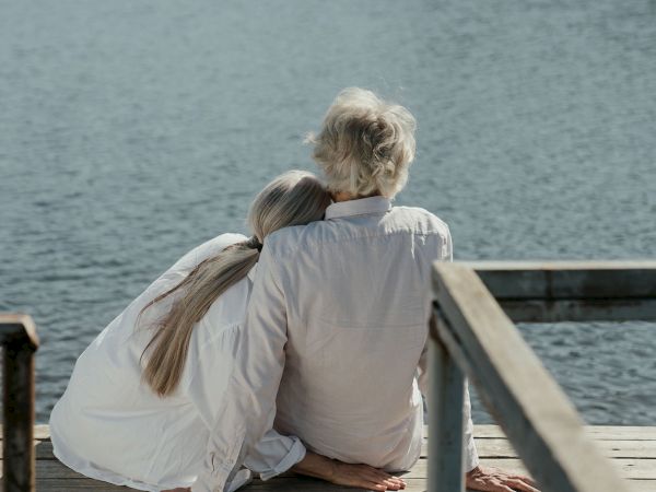 A couple in white shirts sits on a wooden dock hugging while looking out at the ocean.
