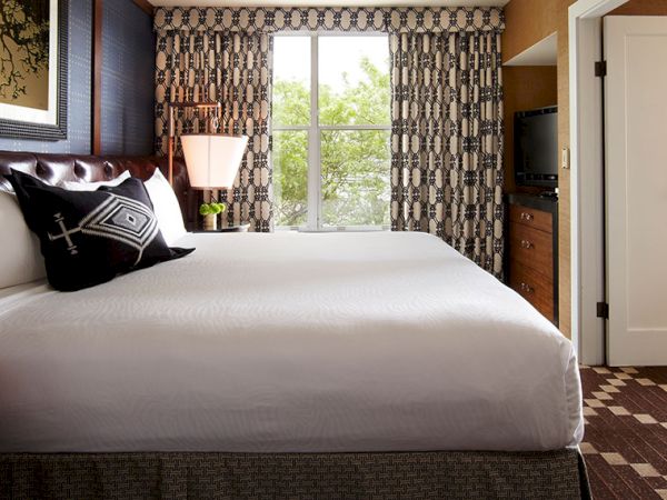 A stylish hotel bedroom with a large bed, patterned curtains, a window, a TV, and a dresser, with a doorway leading out.
