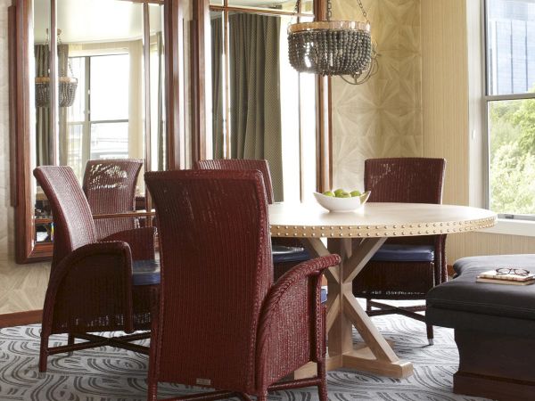 A bright dining room with patterned chairs, a round studded table, hanging beaded chandelier, and large windows with curtains.