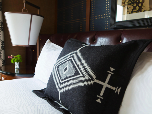 A hotel bed with white bedding and a patterned black-and-white throw pillow, plus a nearby bedside lamp in warm lighting.