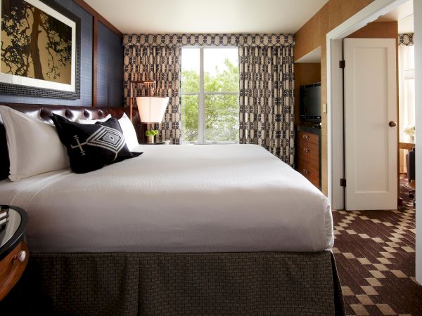 A hotel bedroom with a large bed, patterned curtains, a bedside lamp, and an open doorway to another room.