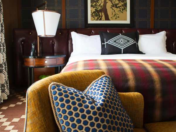 A cozy hotel-style bedroom with a neatly made bed, patterned blanket and pillows, a floor lamp, and framed wall art.