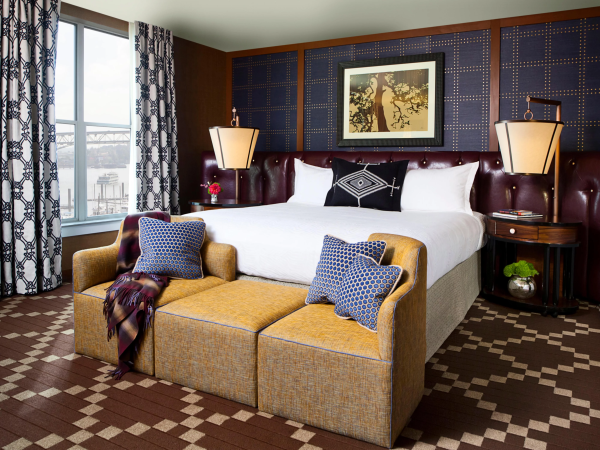 A stylish hotel room with a large bed, patterned curtains, two hanging lamps, framed wall art, and a cushioned seating area by the window.