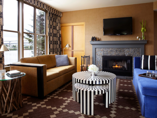 A cozy living room with a couch by big windows, a lit fireplace, a wall-mounted TV, and patterned ottoman tables.