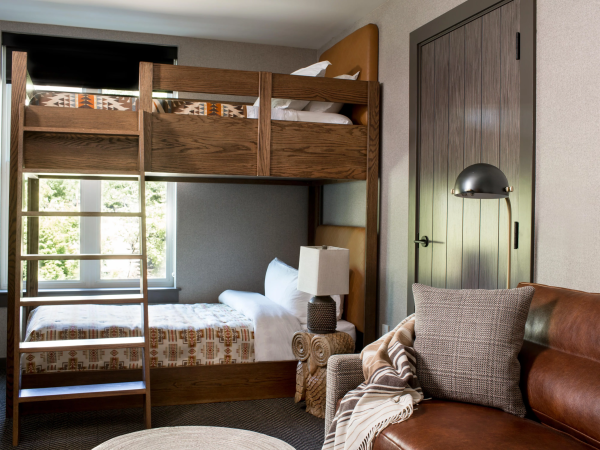 A cozy hotel room with wooden bunk beds, patterned bedding, a sofa by a lamp, and a door with vertical panels.