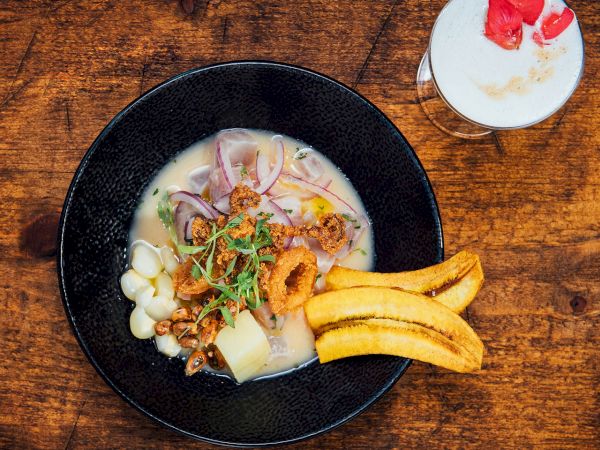 A black bowl of creamy soup with toppings, banana slices, and a small glass of latte topped with strawberry pieces.
