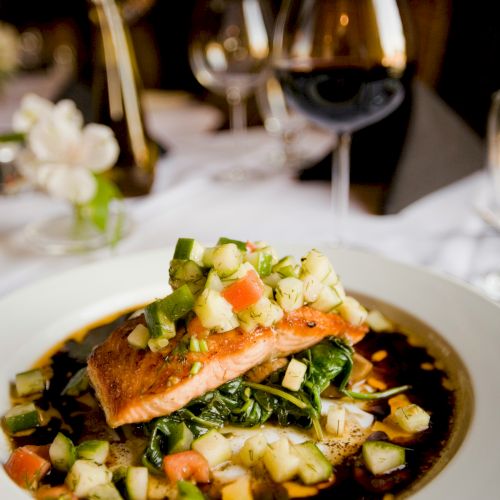 A plated salmon with chopped cucumber, tomatoes, herbs, and greens, with wine glasses and a candlelit dining table in the background.