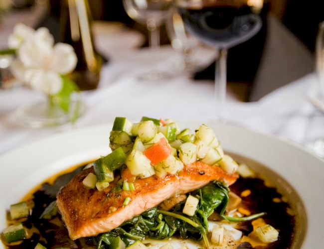 A plated salmon with chopped cucumber, tomatoes, herbs, and greens, with wine glasses and a candlelit dining table in the background.