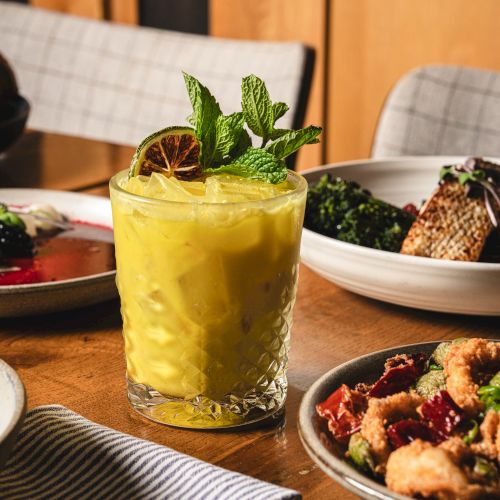 A yellow mint-garnished cocktail in a faceted rocks glass, with plates of food like salads and fries/bites around it.