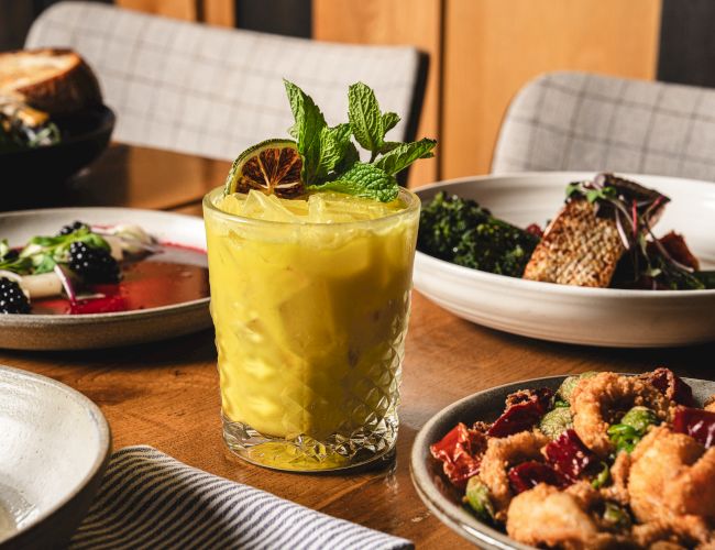 A yellow mint-garnished cocktail in a faceted rocks glass, with plates of food like salads and fries/bites around it.