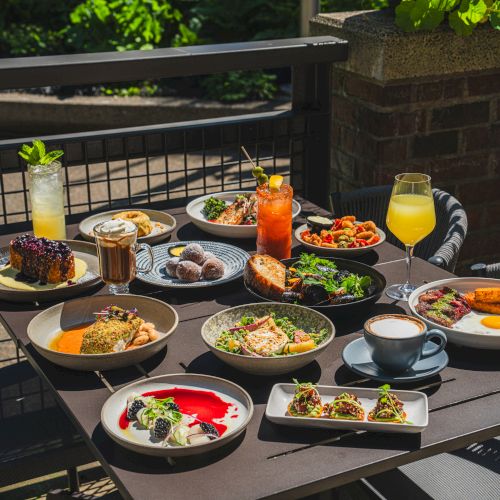 An outdoor patio table is packed with brunch dishes, desserts, and drinks like iced beverages and juice glasses, ready to serve guests.
