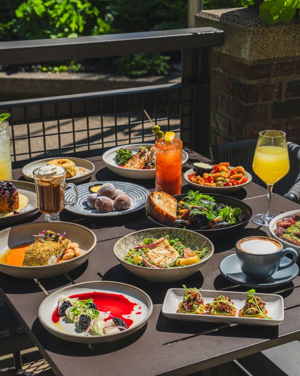 An outdoor patio table is packed with brunch dishes, desserts, and drinks like iced beverages and juice glasses, ready to serve guests.