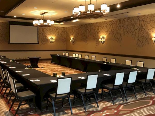 A modern conference room with long tables, chairs, a projector screen, and chandeliers, ready for meetings.