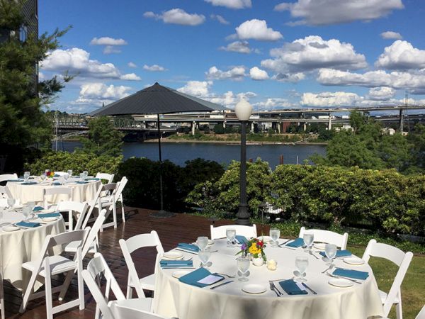 Outdoor dining setup with white chairs and tables overlooking a river, trees, and a bridge under a sunny blue sky.