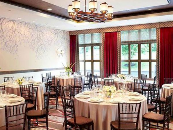 A decorated banquet hall with round dining tables, black chairs, chandeliers overhead, and red-and-gold curtains by tall windows.