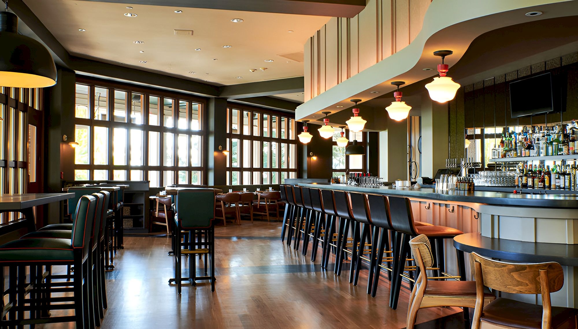 A modern restaurant/bar interior with a long counter, stools, hanging pendant lights, large windows, and rows of bottled drinks.