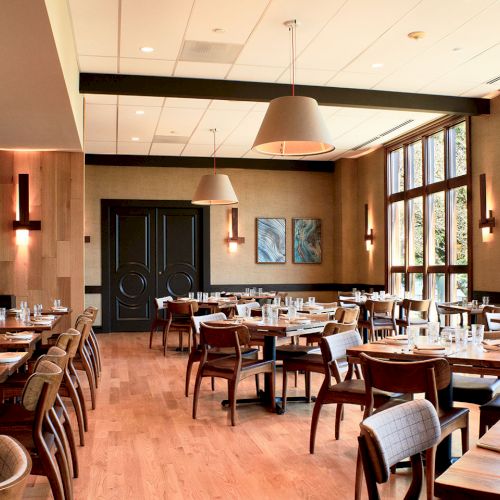 A bright modern restaurant dining room with wooden chairs, neatly set tables, pendant lights, and large windows on both sides.