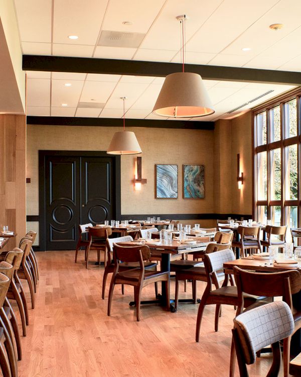 A bright modern restaurant dining room with wooden chairs, neatly set tables, pendant lights, and large windows on both sides.