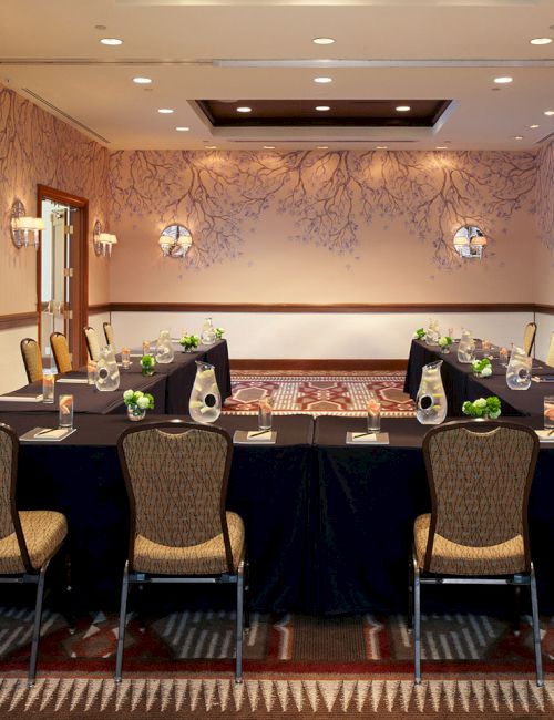 A hotel banquet room set for a formal dinner, with two long tables, chairs, floral centerpieces, and wall sconces.