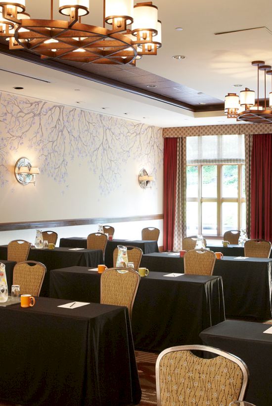 A banquet meeting room set with round tables, chairs, cups, and pitchers under warm chandeliers and patterned wall panels.