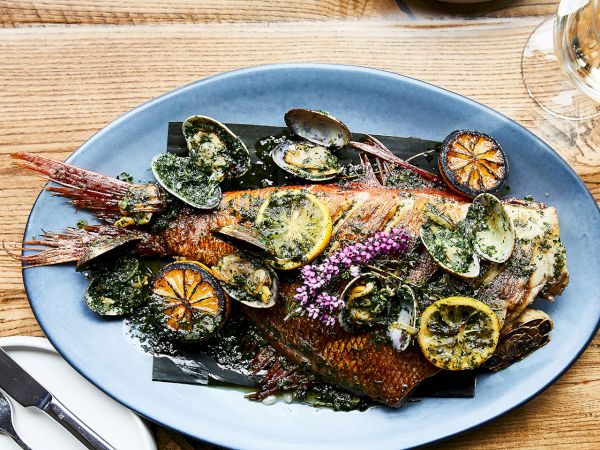 A blue plate of grilled fish topped with lemon slices, mussels, herbs, and edible flowers, with wine nearby.