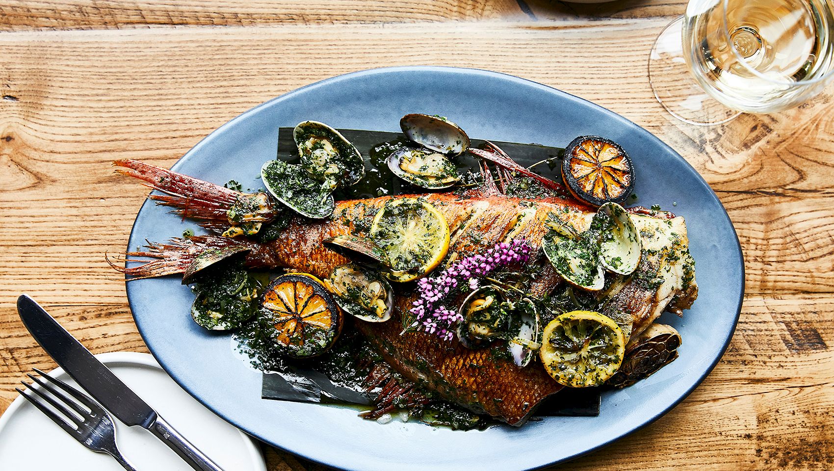 A blue plate holds grilled fish topped with mussels, lemon slices, herbs, and flowers, beside a glass of white wine.