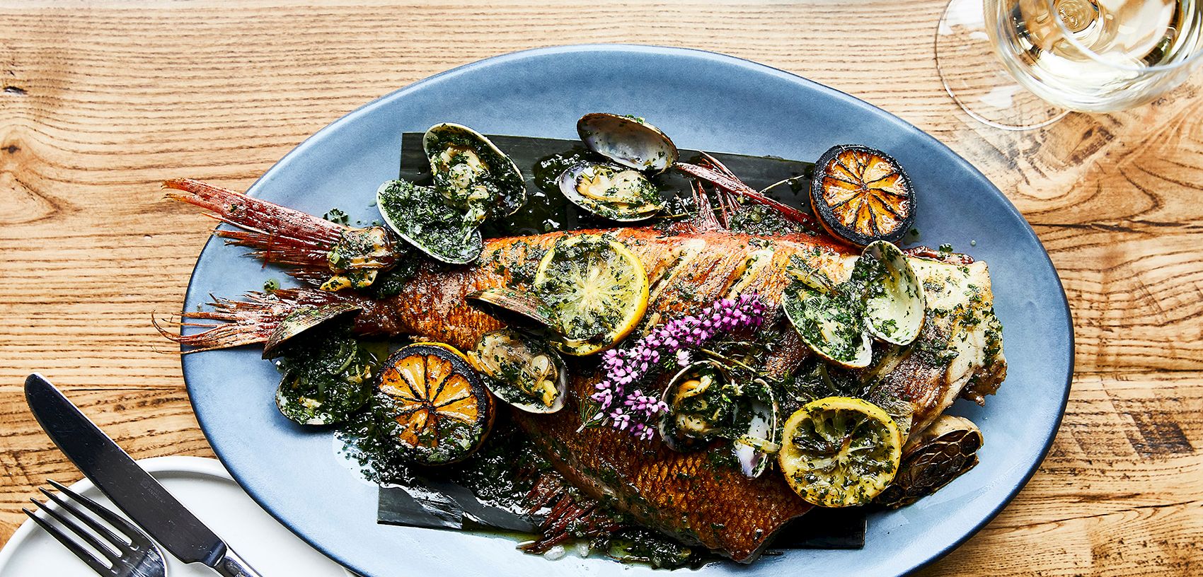 A blue plate holds grilled fish topped with mussels, lemon slices, herbs, and flowers, beside a glass of white wine.