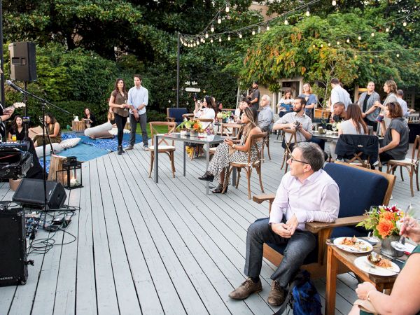 An outdoor event with a DJ/keyboard on stage, a microphone, string lights, and guests seated around tables.