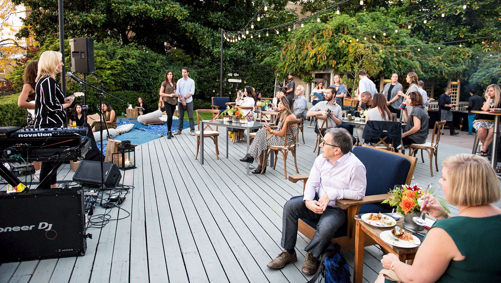 Outdoor patio event with a live DJ onstage, strings of lights, and guests seated at tables listening to music.