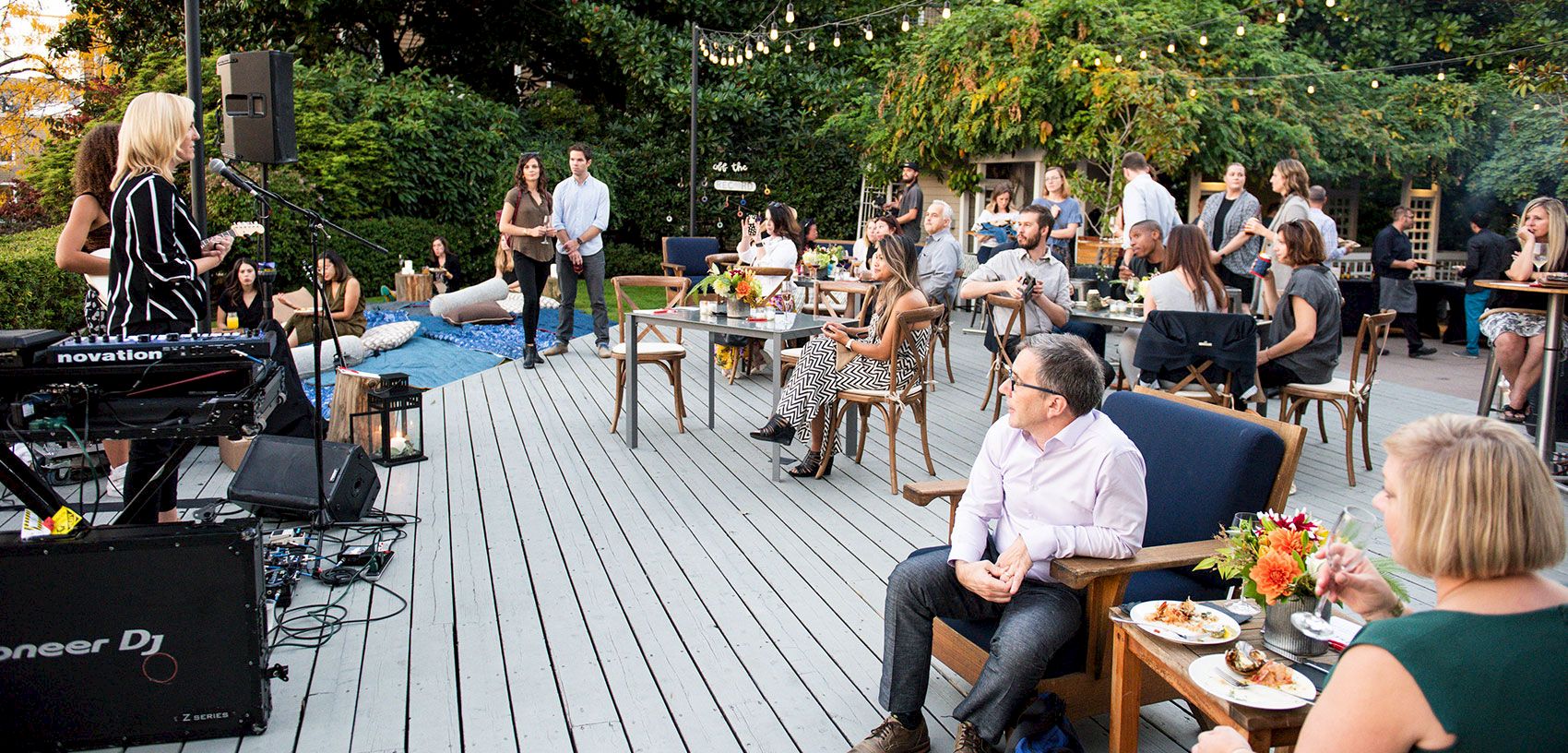 Outdoor patio event with a live DJ onstage, strings of lights, and guests seated at tables listening to music.