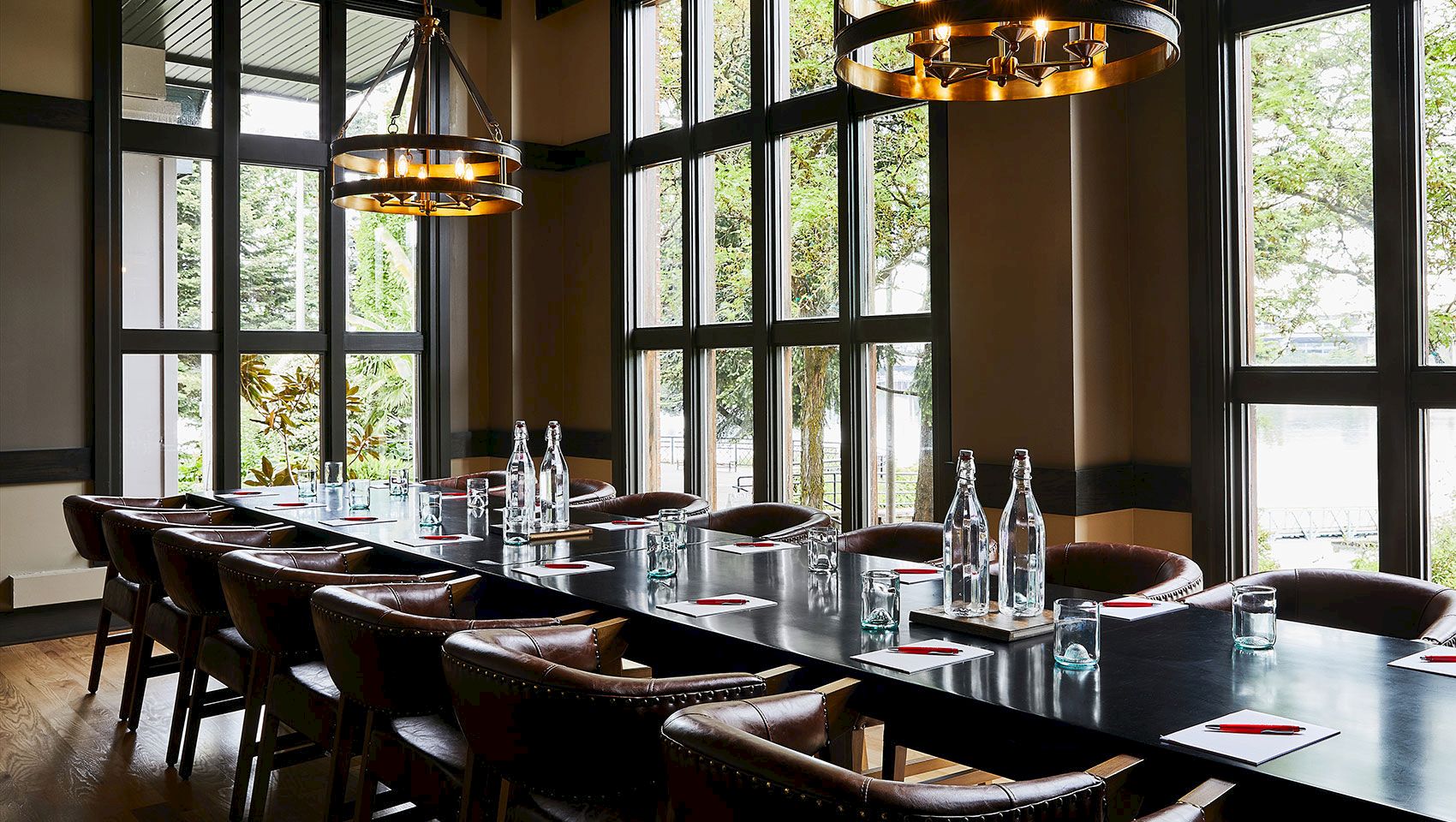 A modern dining room with a long banquet table, leather chairs, bottles and glassware, pendant lights, and large windows overlooking greenery.