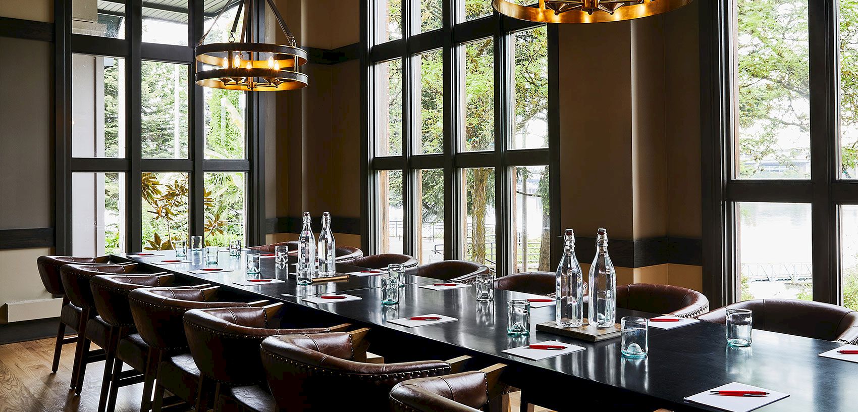 A modern dining room with a long banquet table, leather chairs, bottles and glassware, pendant lights, and large windows overlooking greenery.
