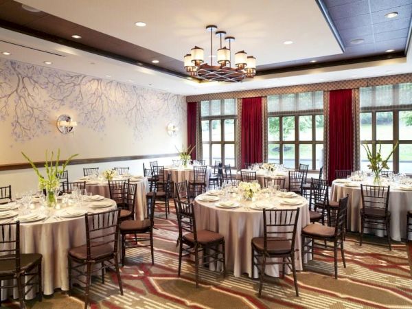 A formal banquet hall set with round tables, white linens, black chairs, red curtains, and a chandelier overhead.