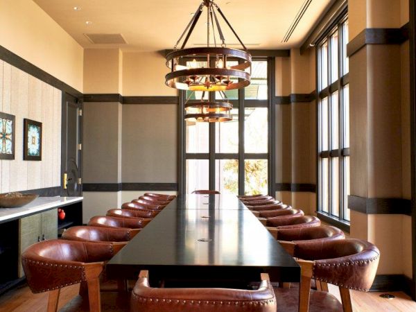 A modern conference room with a large chandelier, a long wooden table, and rows of brown leather chairs.