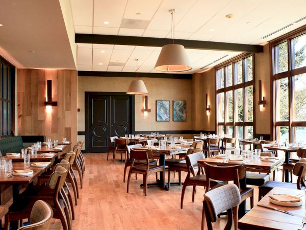 A modern restaurant dining room with wooden tables and chairs, pendant lights, and large windows letting in daylight.