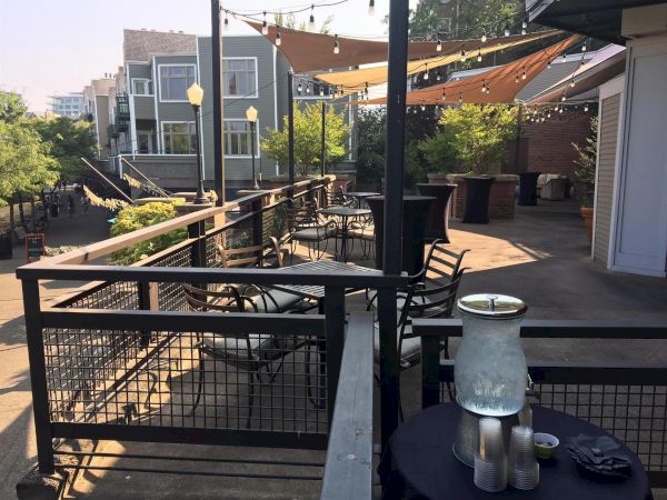 Outdoor patio deck with string lights, tables, chairs, and trees in warm sunlight overlooking buildings.