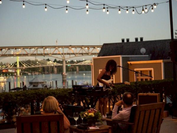 An outdoor night concert: a guitarist performs on a patio with string lights above and guests seated in front of waterfront lights.
