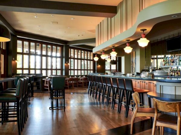 A modern hotel bar interior with wooden stools, bright pendant lights, large windows, and a stocked bottles shelf.