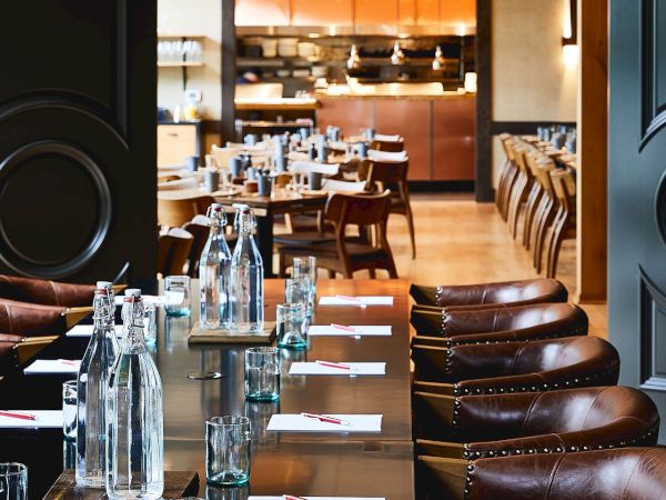 A cozy restaurant dining room with leather chairs, set tables, and glass bottles waiting on a central table.