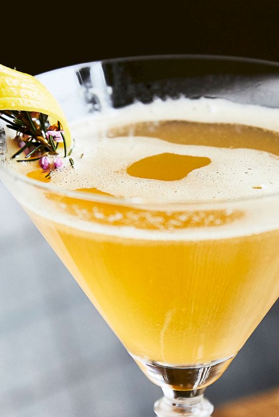 A golden cocktail in a martini glass with foam, garnished with a yellow citrus twist and tiny purple flowers, ready to sip.